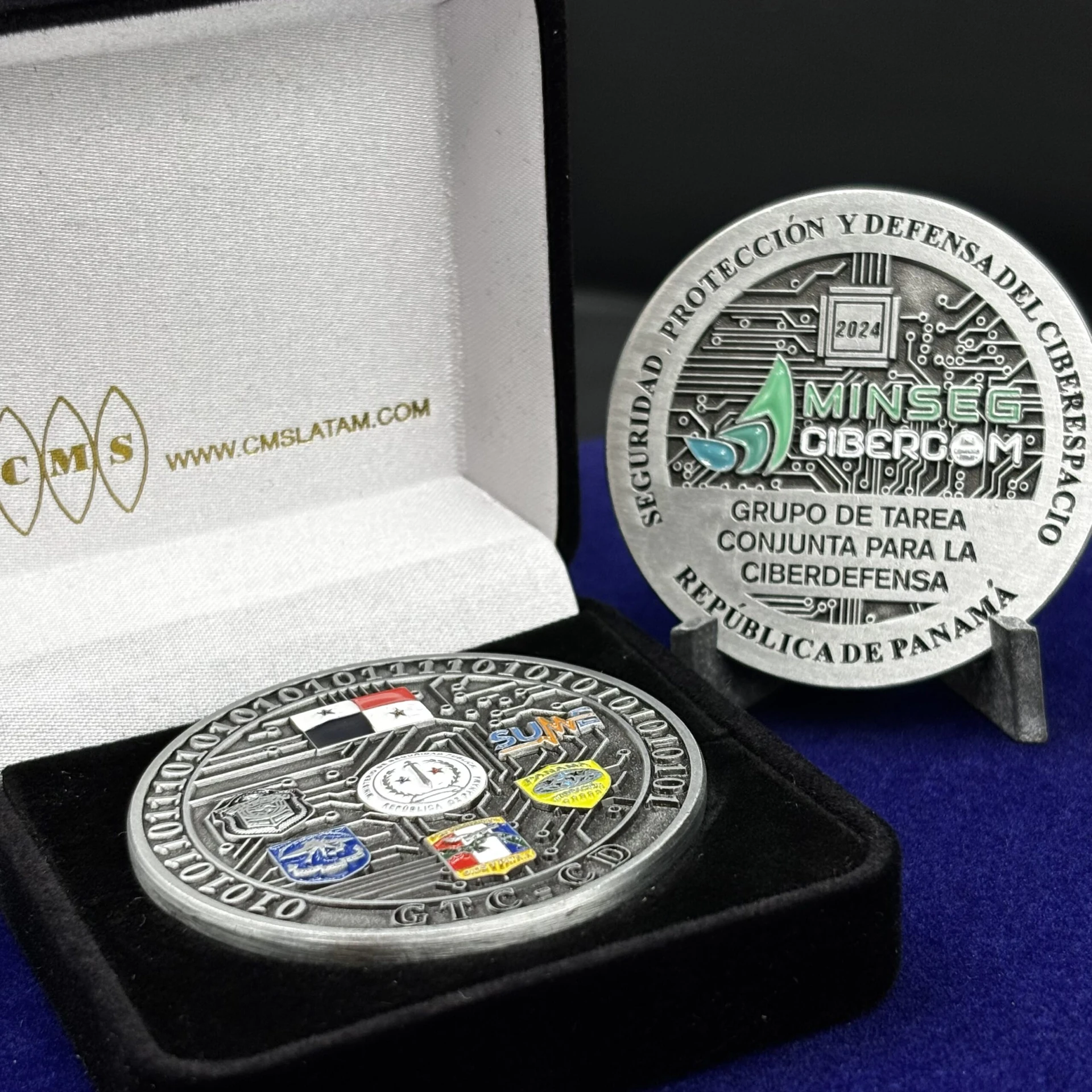 Moneda de ciberdefensa por ambas caras, con relieve plateado y detalles de color, junto a un estuche CMS.